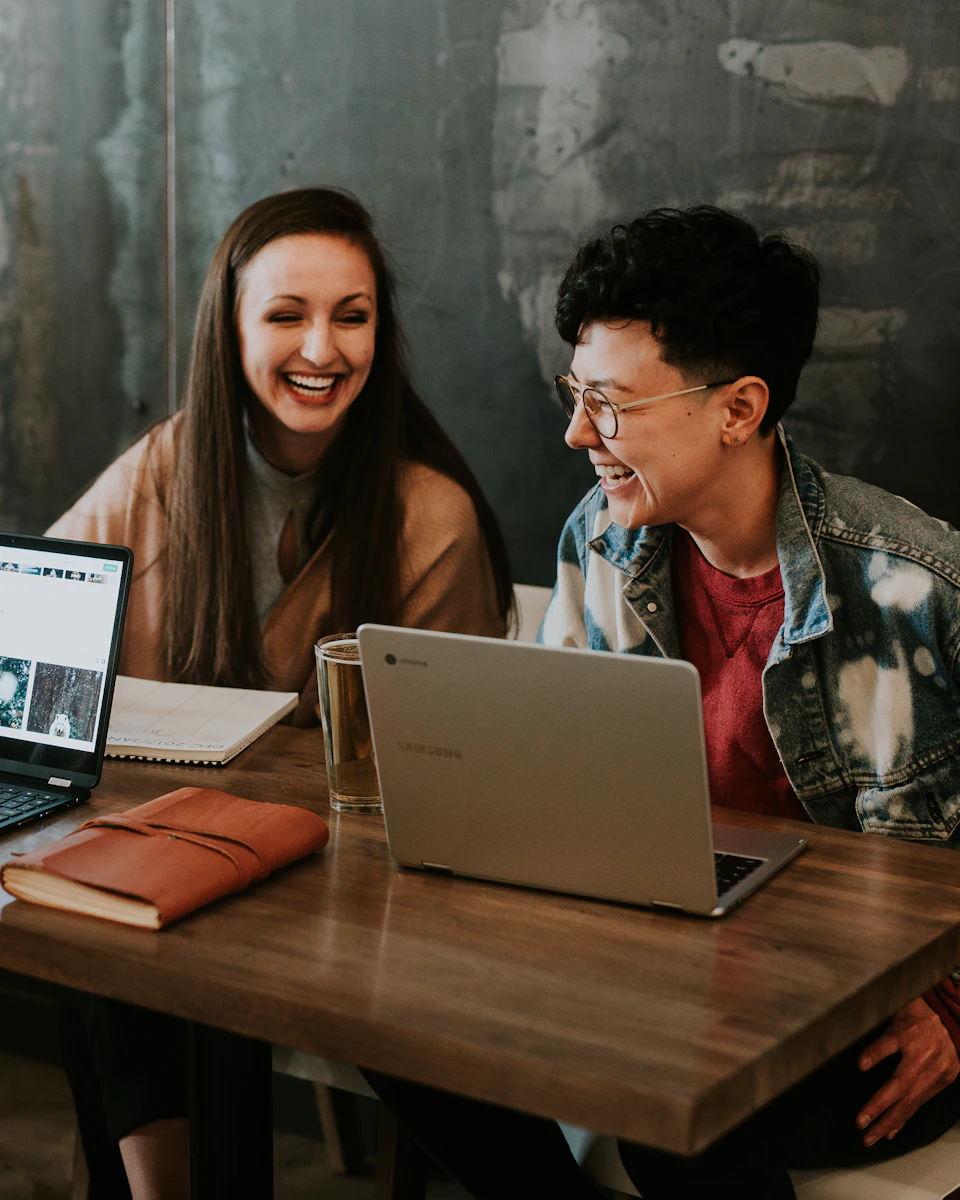 Two colleagues laughing together at a table with laptops and a notebook